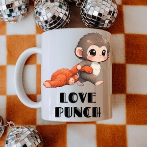 May include: White ceramic mug featuring a cartoon illustration of two monkeys. One monkey is lying down, and the other is standing and holding the other monkey's arm. The mug has the words "LOVE PUNCH" in black block letters.