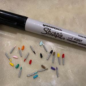 May include: A collection of colorful marker tips, including red, orange, yellow, green, blue, pink, and brown, scattered on a light gray surface. A black and white Sharpie fine point permanent marker is also visible in the image.