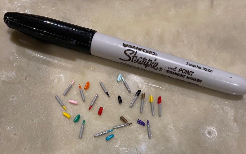 May include: A collection of colorful marker tips, including red, orange, yellow, green, blue, pink, and brown, scattered on a light gray surface. A black and white Sharpie fine point permanent marker is also visible in the image.