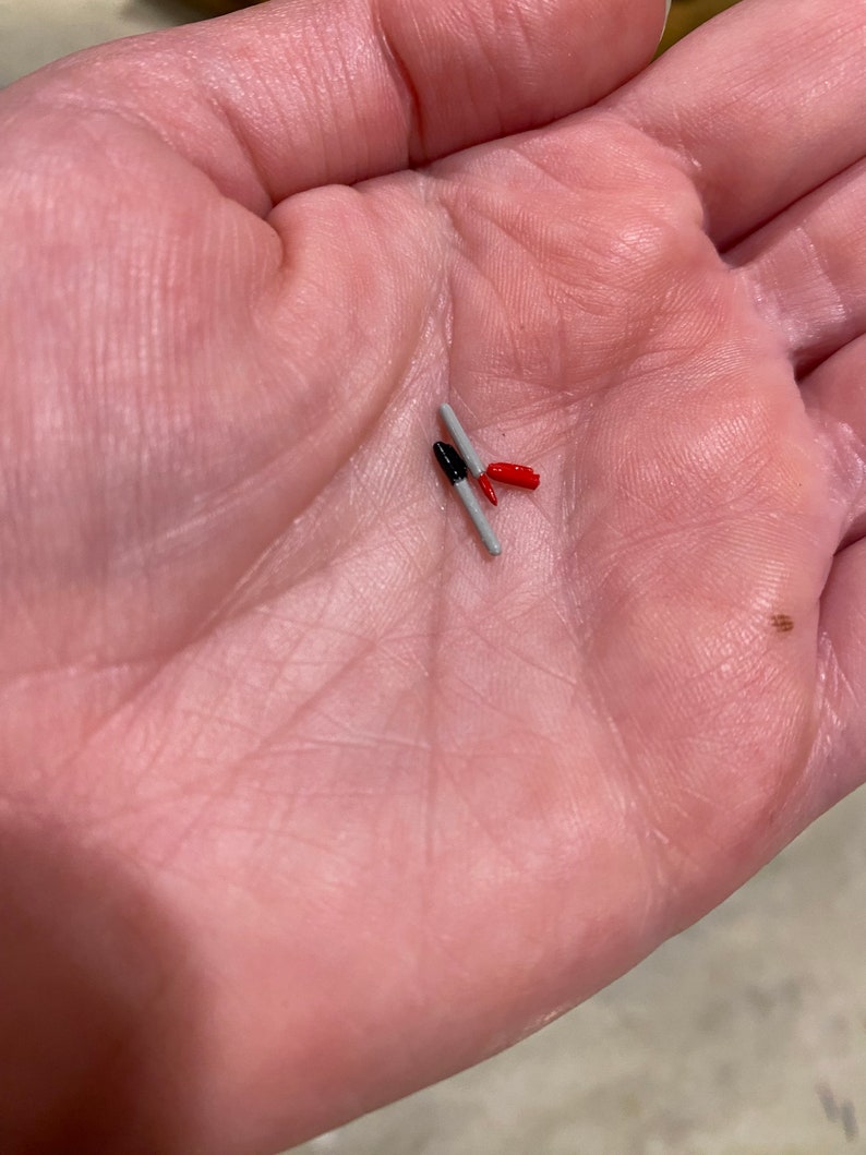 May include: Two small, gray, cylindrical objects with red and black tips. The objects are lying on a person's palm.