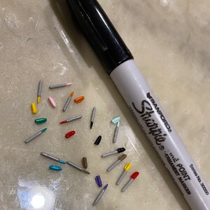 May include: A close-up of a variety of colorful markers with silver caps. The markers are scattered on a light gray surface. A black Sharpie permanent marker is also visible in the image.