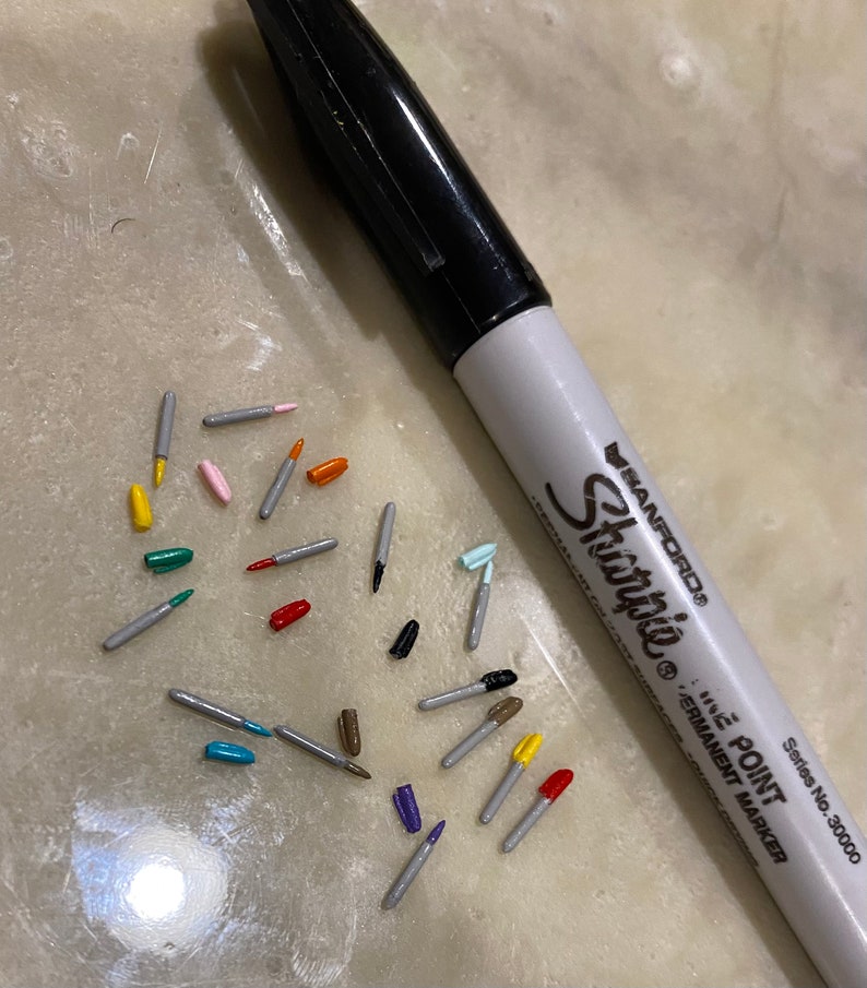 May include: A close-up of a variety of colorful markers with silver caps. The markers are scattered on a light gray surface. A black Sharpie permanent marker is also visible in the image.