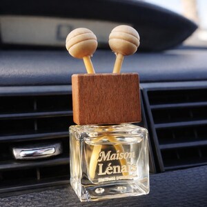 Könnte beinhalten: Auto-Lufterfrischer mit einer klaren, quadratischen Glasflasche, die eine hellfarbene Flüssigkeit enthält. Die Flasche wird von einem Holzblock mit zwei hellfarbigen, kugelförmigen Holzdiffusoren gekrönt. Der Text "Maison Lénais" ist sichtbar.
