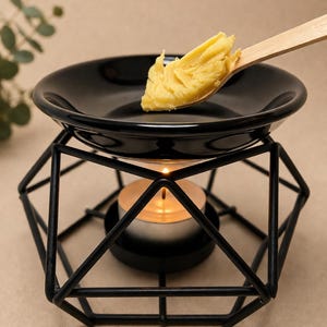 May include: A black ceramic wax warmer with a geometric metal frame. A lit tea light candle sits inside the frame, beneath the warmer's dish. A wooden spoon holds a portion of yellow wax, ready to melt.