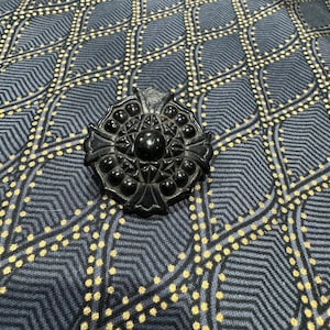 May include: A black, ornate, circular brooch with a raised, geometric design. The brooch has a central, circular, raised, black stone.