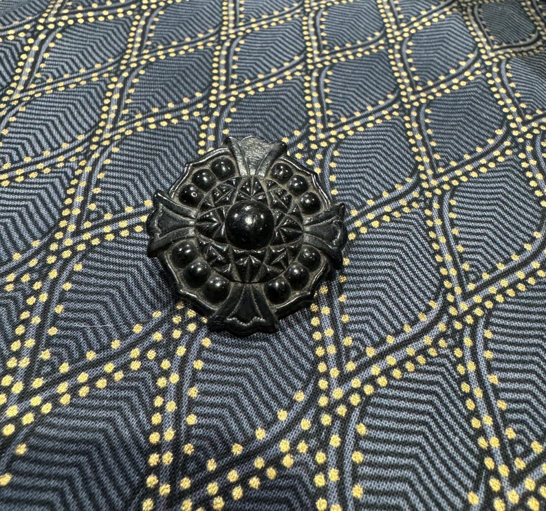 May include: A black, ornate, circular brooch with a raised, geometric design. The brooch has a central, circular, raised, black stone.