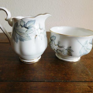 May include: A white porcelain creamer and sugar bowl set. The set features a delicate leaf pattern in shades of blue and gray, with gold trim. The creamer has a handle and a spout. The sugar bowl is open and round.