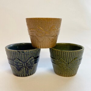 Peut inclure: Trois pots en céramique de couleurs marron, bleu marine et vert. Chaque pot présente un motif côtelé et un détail de nœud. Les pots sont empilés, mettant en valeur leur design décoratif et fonctionnel.