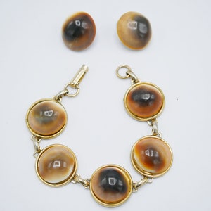 May include: A gold bracelet with five round brown and white shell-like beads. The bracelet is paired with two matching earrings.