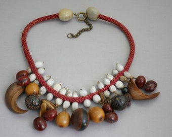 May include: A handcrafted necklace featuring a braided red cord with various beads and charms. The necklace includes white, brown, and black beads, along with wooden and amber-colored charms. The necklace has a brass clasp and chain.