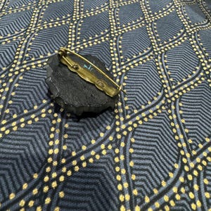 May include: A black and gold brooch with a textured, irregular shape. The brooch has a pin and catch closure.