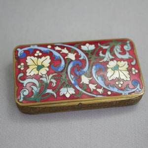 May include: Rectangular trinket box with a red background and floral design. The box features a gold-colored frame and is adorned with white, yellow, blue, and green floral patterns. The box is likely made of metal and has a hinged lid.