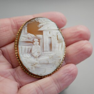Pu&ograve; includere: Un broche in cammeo bianco con una montatura dorata. Il cammeo presenta una scena intagliata di una donna in piedi davanti a un edificio.