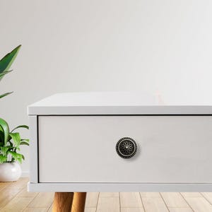 Könnte beinhalten: Eine weiße Schublade mit einem dekorativen schwarzen und goldenen Blumenknauf. Der runde Knauf hat ein detailliertes Blumendesign und einen gepunkteten Rand. Die Schublade ist Teil eines weißen Schranks mit Holzbeinen.