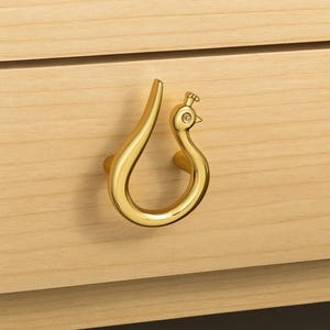 May include: A gold-colored peacock-shaped drawer pull mounted on a light wood drawer. The pull features a curved handle and a detailed peacock head with a small gem for the eye. The drawer is made of light-colored wood.