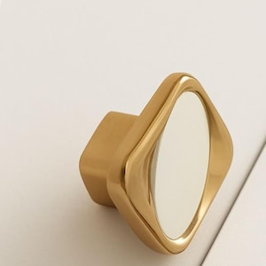 Könnte beinhalten: Ein goldfarbener Schrankknauf mit rautenförmiger Front und quadratischer Basis. Der Knauf hat eine glatte, reflektierende Oberfläche und ein modernes Design. Geeignet für Schubladen und Schränke.