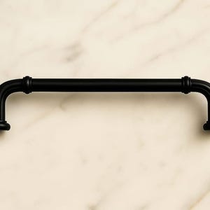 Puede incluir: Un tirador de armario negro mate con una barra tubular y extremos curvos. El herraje tiene un diseño simple e industrial, con una barra cilíndrica y pies redondeados. El tirador está sobre un fondo claro, similar al mármol.
