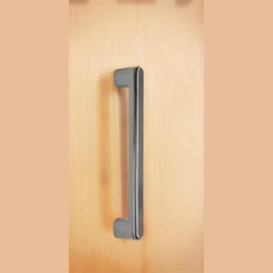 Könnte beinhalten: Ein polierter Türgriff aus Chrom mit rechteckigem Design. Der Griff ist an einer hellen Holztür befestigt. Der Griff ist etwa 30 cm lang.