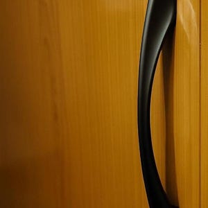 Puede incluir: Primer plano de una puerta de armario de madera con un tirador negro curvado. La puerta tiene un color marrón dorado cálido, y el tirador es elegante y moderno, proporcionando un contraste con la veta de la madera.