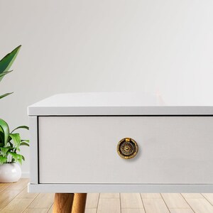 May include: A gold-colored drawer pull with a floral design is attached to a white drawer. The drawer is part of a white cabinet with wooden legs. A green plant in a white pot is in the background.