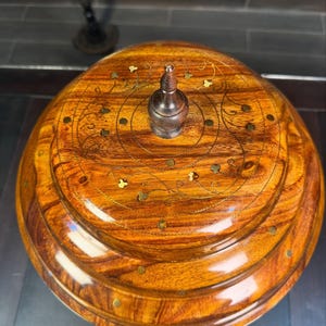 May include: A round, polished wooden container with a tiered lid featuring a central dark wooden finial. The surface displays a natural wood grain pattern, accented with inlaid brass dots and delicate, etched floral vine motifs.