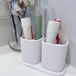 May include: A white, ribbed toothbrush holder with two compartments, holding a green electric toothbrush, two tubes of toothpaste, and a red toothbrush. The holder sits on a white surface, with a glass vase of cotton stems in the background.