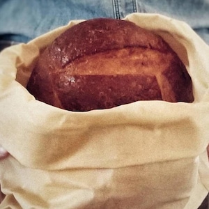 May include: A freshly baked loaf of bread, golden brown with scored top, nestled in a beige cloth bag. The bread has a glossy crust and is presented in a soft, natural-toned bag, suggesting a rustic or artisanal bakery.