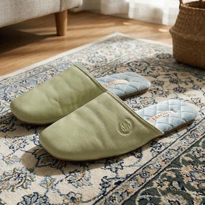 May include: A pair of sage green leather slippers with a quilted light blue interior. The slippers have a square toe design and a small embossed emblem. The slippers are resting on a patterned rug.
