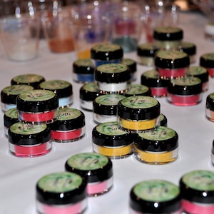 May include: A collection of small, round, glass jars with lids. The jars are filled with colorful pigments and powders. The jars are arranged in a grid pattern on a white surface.