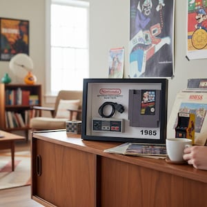 Pode incluir: Uma exibição emoldurada do sistema de entretenimento Nintendo com um cartucho de jogo, um controlador e um cabo. A exibição inclui o texto "1985". A cena inclui cartazes vintage e um armário de madeira.
