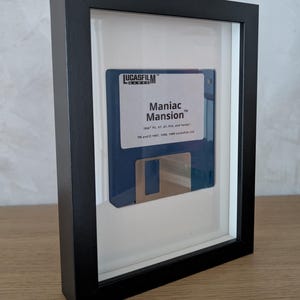 Könnte beinhalten: Gerahmte Vintage-Diskette von Maniac Mansion. Die blaue Diskette wird in einem schwarzen Rahmen mit weißem Passepartout präsentiert. Der Text "Maniac Mansion" ist auf der Diskette zusammen mit dem Lucasfilm Games-Logo zu sehen.