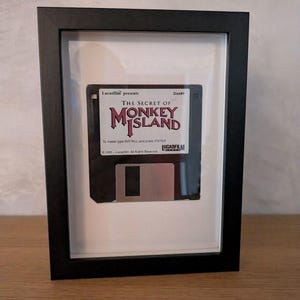 Puede incluir: Disquete negro enmarcado con el texto "THE SECRET OF MONKEY ISLAND" y "Lucasfilm presents." El disco está montado sobre un fondo blanco dentro de un marco negro, creando una estética vintage.