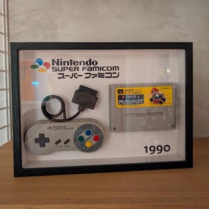 Könnte beinhalten: Gerahmte Nintendo Super Famicom-Anzeige. Die Anzeige enthält einen grauen Controller, eine Spielkassette und den Text "Nintendo SUPER FAMICOM" und "1990". Die Spielkassette zeigt den Text "MARI KART".