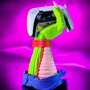 May include: A green hand-shaped controller stand holding a white and black gaming controller. The base is blue with a pink textured conical section and a red band. The stand features sharp white claws and a vibrant, multi-coloured design.