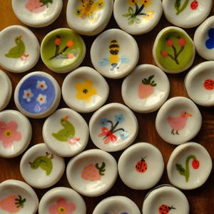 Op de afbeelding: Een verzameling kleine, witte keramische schaaltjes, elk met de hand beschilderd met kleurrijke ontwerpen van insecten, bloemen en fruit. De ontwerpen omvatten bijen, vlinders, aardbeien en lieveheersbeestjes. Deze decoratieve schaaltjes zijn ideaal.