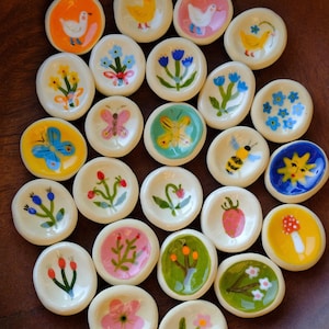 May include: A collection of small, round, off-white ceramic dishes, each featuring a unique hand-painted design including flowers, butterflies, bees, birds, a sun, and a mushroom in vibrant colours like orange, blue, pink, yellow, and green.
