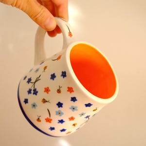 以下が含まれることがあります： 青とオレンジの花のフローラルパターンと青い縁が付いた白いセラミックマグ。マグの内側は明るいオレンジ色です。マグは手で持たれています。