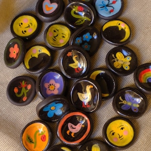 Puede incluir: Una colección de pequeñas piezas redondas de cerámica marrón oscuro, cada una pintada a mano con diseños caprichosos. Los diseños incluyen flores, lunas, caras, mariposas y pájaros en colores brillantes como amarillo, azul y naranja. Las piezas están dispuestas sobre una superficie de tela neutra.
