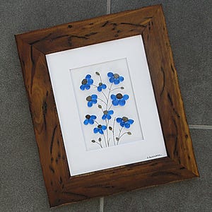 May include: A framed pebble art piece featuring blue flower designs. The artwork is set against a white background within a rustic brown wooden frame. The text "Pebble Art by D. Dellatore" and "Winterberry Originals" are visible.