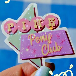 Könnte beinhalten: Ein holografischer Aufkleber mit einem Schild-Design im Retro-Stil. Der obere Teil zeigt das Wort PINK in gelben Buchstaben auf rosa Kreisen. Darunter steht auf einem violetten rechteckigen Schild Pony Club in gelber Schreibschrift mit Sternen.