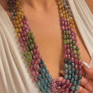 Puede incluir: Un collar de múltiples hilos con cuentas pulidas de forma ovalada en un degradado de colores que incluye amarillo, verde, rosa y azul. Las cuentas están ensartadas en un hilo oscuro, creando un efecto de capas. Un logotipo con un arcoíris y el texto KUNDAN ERA STUDIO es visible.