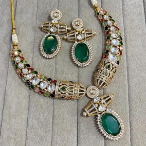Puede incluir: Un conjunto de collar y pendientes dorados. El collar y los pendientes presentan piedras verdes esmeralda de corte ovalado, rodeadas de piedras preciosas claras y coloridas. La joyería está sobre un fondo neutro.