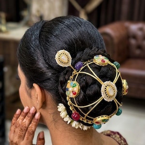 May include: A gold-toned metal hair accessory featuring a spherical cage design adorned with multi-coloured cabochon stones and white floral accents. The piece is secured over a dark, braided hair bun, showcasing intricate filigree details and beads.
