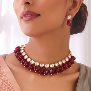 May include: A gold-toned necklace and matching earrings set. The necklace features a series of dark red, teardrop-shaped beads, interspersed with clear, rectangular stones. The earrings have a similar design, with a single red bead.