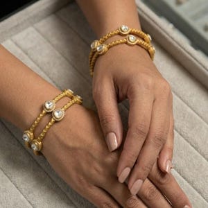 May include: Close-up of two gold bangles with clear, round gemstones. The bangles have a textured, braided design and are worn on a person's wrists. The background is a soft gray surface.