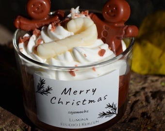 Puede incluir: Un recipiente de vela de cristal relleno de cera marrón, cubierto con cera batida blanca, dos figuras de hombre de jengibre marrones y una forma de bastón de caramelo blanco. Una etiqueta blanca dice Merry Christmas, sojawachs, LUMINA STUDIO | KERZEN.