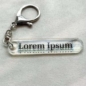 May include: A clear rectangular keychain with the text "Lorem Ipsum" printed in black. The keychain has a silver-coloured clasp and chain. The background is a light-coloured fabric.