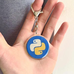 Puede incluir: Un llavero redondo con el logotipo del lenguaje de programación Python en blanco y amarillo sobre un fondo azul. Tiene un cierre plateado y un pequeño lazo para sujetar llaves.