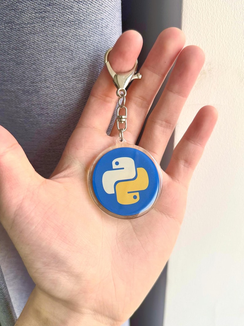 Puede incluir: Un llavero redondo con el logotipo del lenguaje de programaci&oacute;n Python en blanco y amarillo sobre un fondo azul. Tiene un cierre plateado y un peque&ntilde;o lazo para sujetar llaves.
