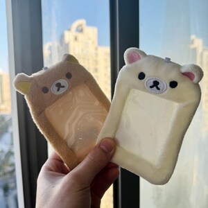 May include: Two plush bear-shaped ID badge holders. One is tan, the other is off-white. Both have a clear plastic window for an ID card, embroidered black eyes, and a small black nose. The ears have pink accents.